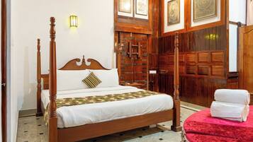 Una cama con dosel y tablero de madera, una mesita de noche, un asiento con cojín rojo y una lámpara fijada a la pared.