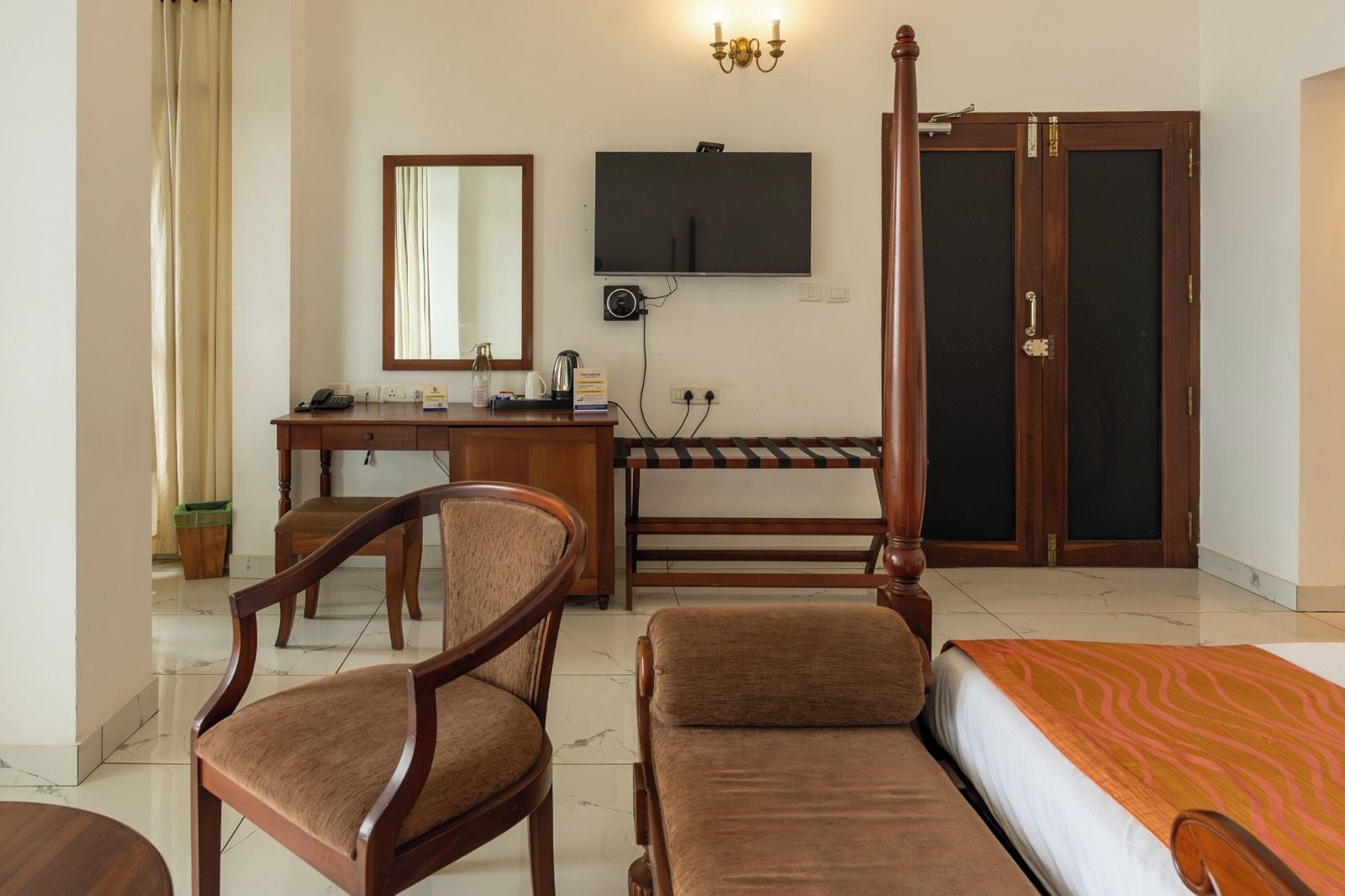A hotel room with a bed, two wooden chairs, a desk with a mirror, a television, and a wardrobe.
