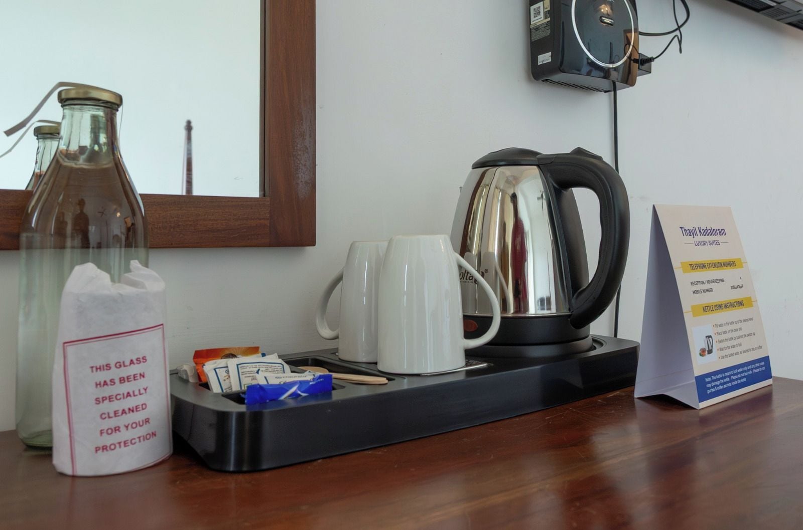 Een hotelkamer met een koffiezetapparaat, een waterkoker, een glazen fles, een mok en een bord met contactgegevens.
