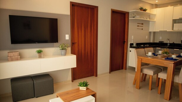 A modern apartment with a flat-screen TV, wooden doors, a dining area with a table and chairs, and a small white table with a plant. - Apto c Churrasqueira e Piscina a 100m da Praia (Japaratinga)