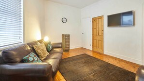 A living room with a leather sofa, patterned cushions, a dark rug, wooden flooring, a clock on the wall, and a door with a window. - Host Stay The Reading Room (Hexham)