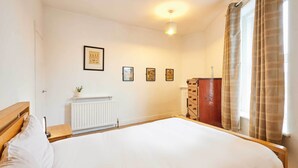 A bedroom with a large bed, a radiator, a wooden nightstand, a dresser, and a window with curtains. - Host Stay The Reading Room (Hexham)