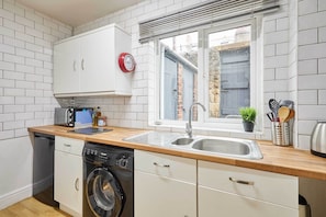A compact kitchen with white cabinets, a wooden countertop, a washing machine, a sink, and a window with blinds. - Host Stay The Reading Room (Hexham)