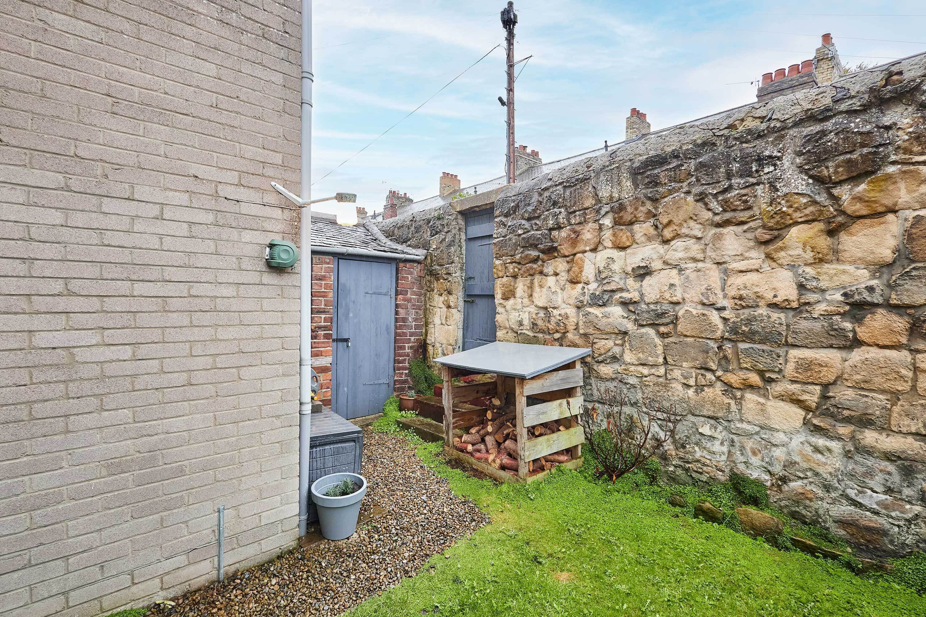 A narrow alleyway with a stone wall on one side and a brick wall on the other. There is a small wooden structure with a metal roof, possibly a shed or storage area, and a grey bucket on the ground.