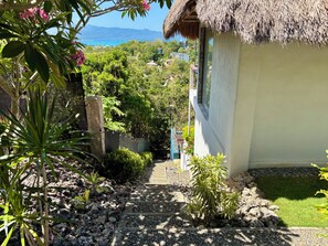 A pathway leading to a house with a thatched roof, surrounded by lush greenery and a view of the sea.