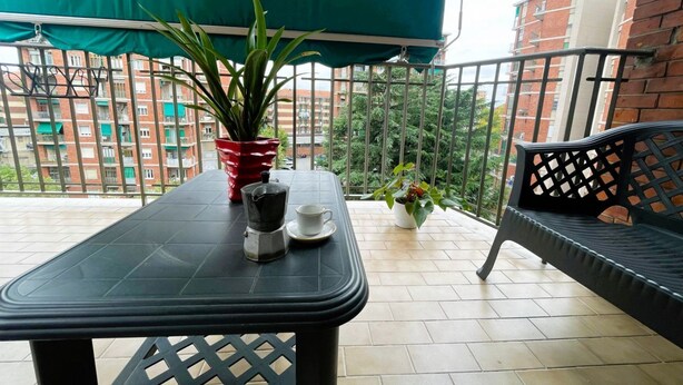 Ein Balkon mit Tisch, einem roten Topf, einem weißen Topf, einer schwarzen Bank und Blick auf Gebäude.