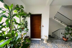 Una entrada de casa con puerta de madera, felpudo, escalera con barandilla metálica y plantas en macetas.