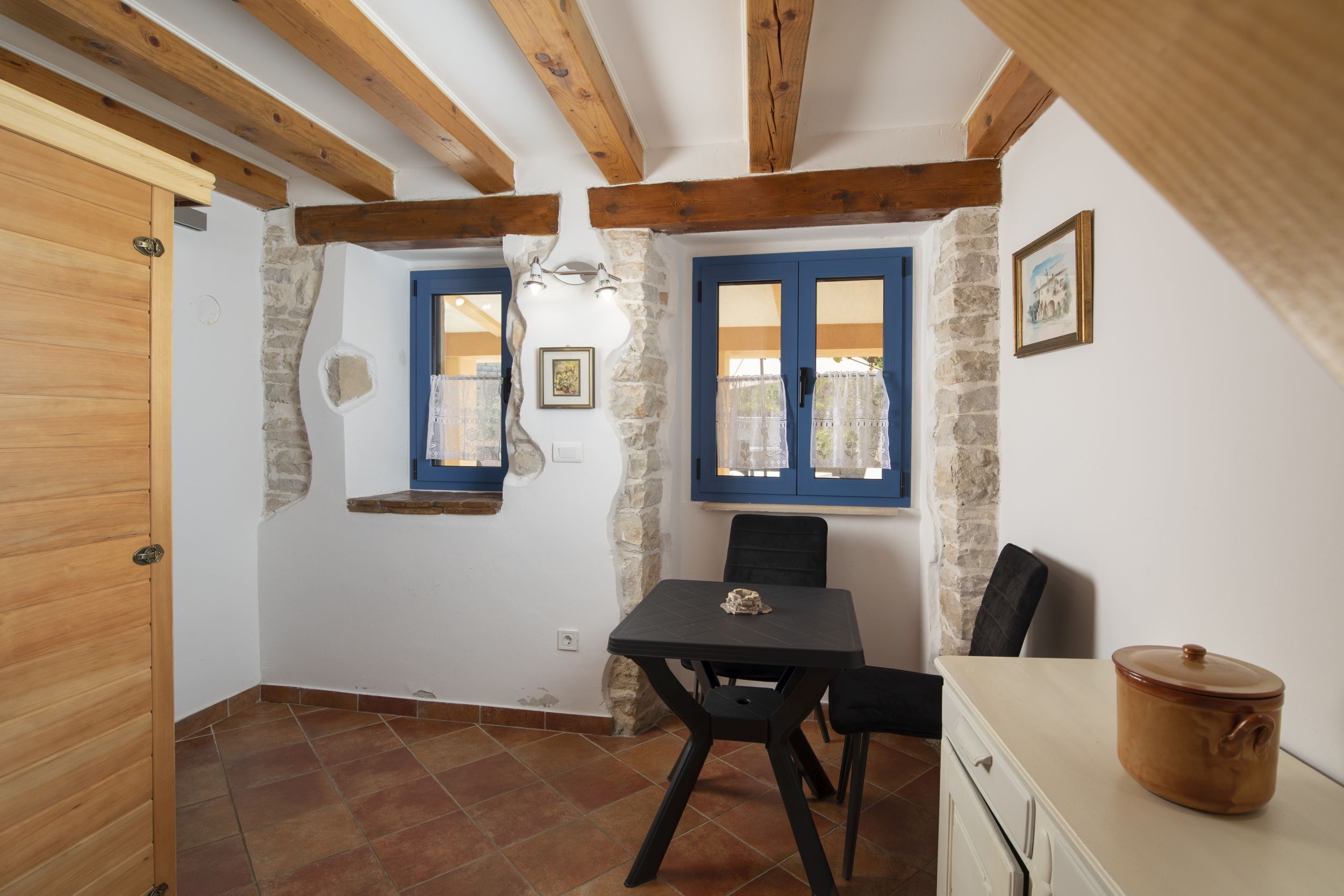 Ein rustikaler Raum mit freiliegenden Holzbalken, einer Steinwand, einem blau gerahmten Fenster, einem Tisch mit Stühlen und einem weißen Schrank mit einem Topf.