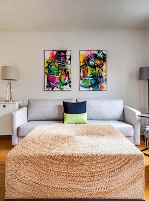 Un salon moderne avec un canapé gris, deux tableaux abstraits, une table basse en osier et une lampe sur une table d’appoint.