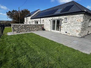 Une maison en pierre avec un toit en pente et des panneaux solaires, une allée pavée et une pelouse bien entretenue.