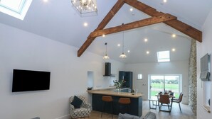 A spacious living area with a high ceiling and exposed wooden beams, a kitchen with a central island, and a dining area with a view of the outdoors. - Beudy Tanrallt (Holyhead)