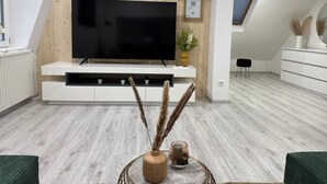 A modern living room with a large flat-screen TV, wooden panel wall, white entertainment unit, and a view of the sky through a skylight.