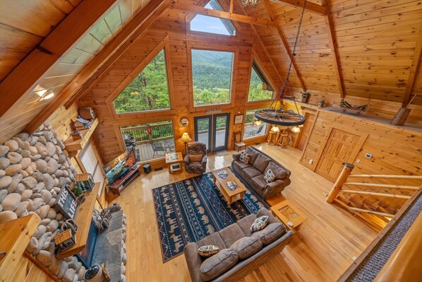 A spacious loft-style cabin with a high ceiling, large windows offering a view of the forest, a cozy seating area with a sofa and coffee table, and a stone wall feature.