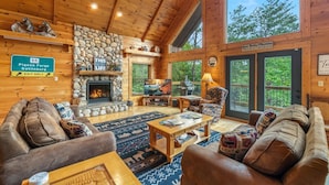 A cozy cabin interior with a stone fireplace, wooden furniture, and a large window overlooking greenery.