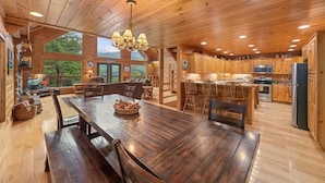 A spacious wooden cabin with a large dining table, a kitchen with wooden cabinets, and a view of the surrounding forest.