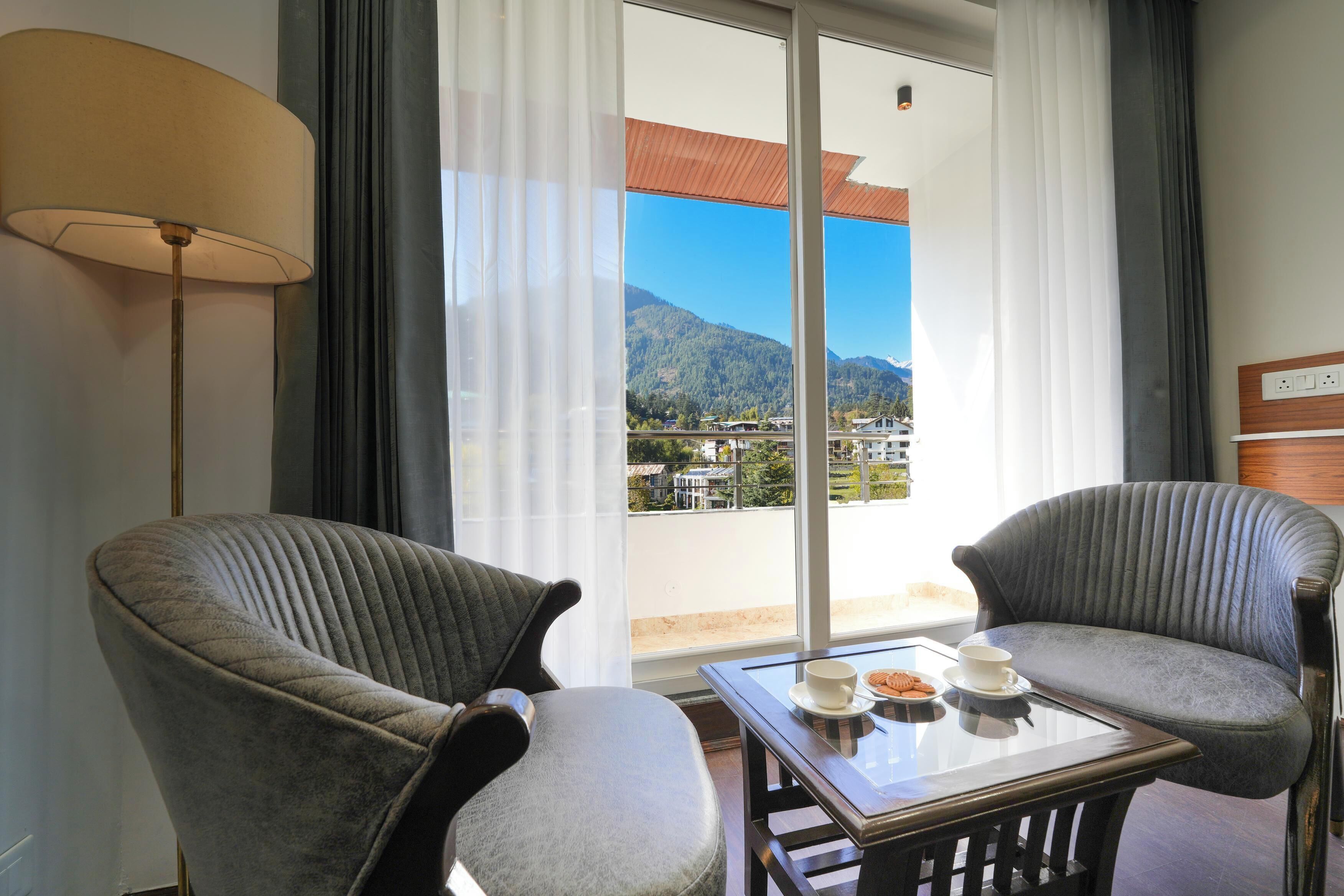 Una habitación con dos sillones, una mesa con una taza y galletas, y vistas a montañas a través de amplios ventanales.
