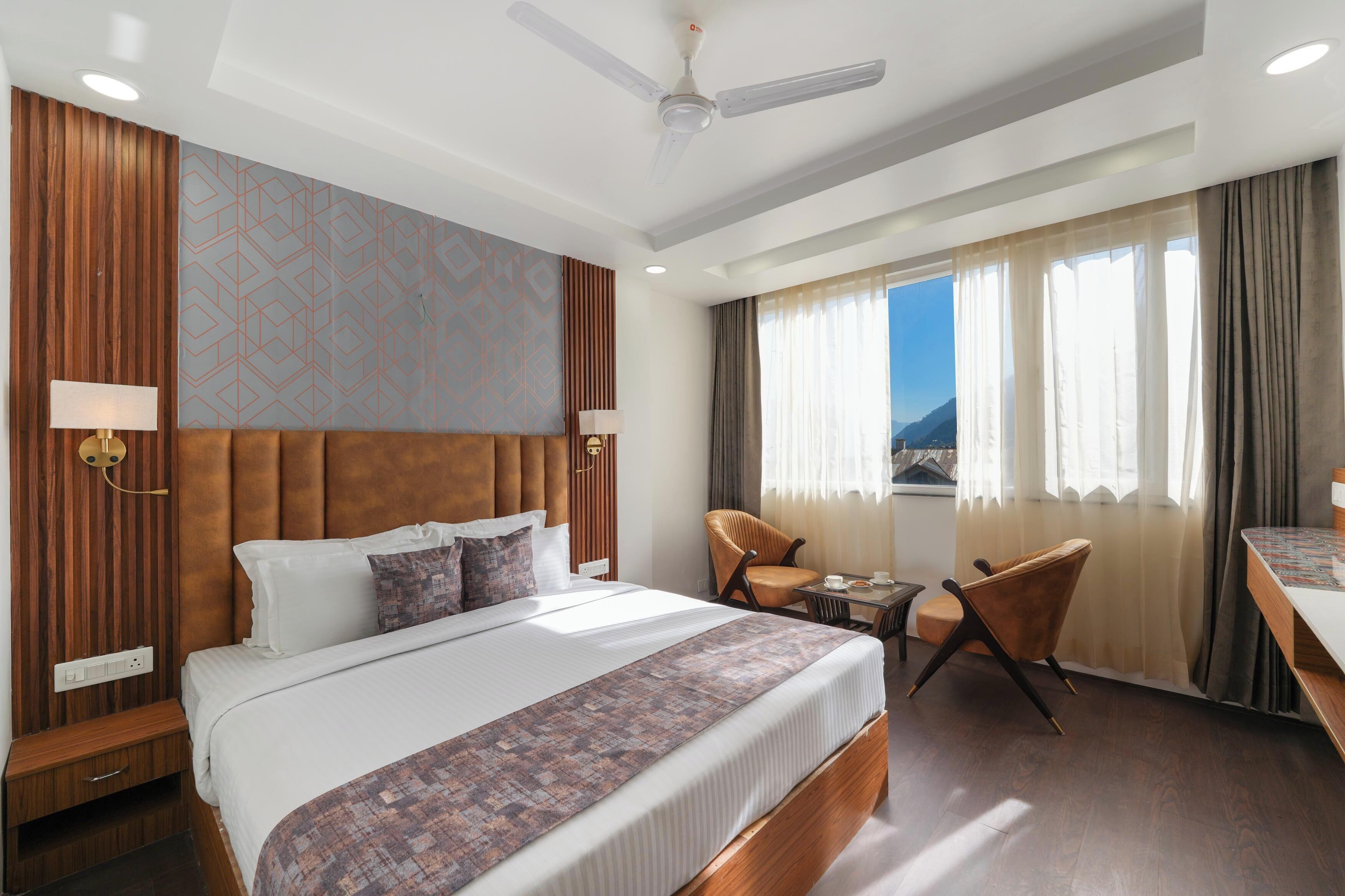 Une chambre d’hôtel avec un grand lit, un bureau, deux chaises, un ventilateur de plafond et une vue sur les montagnes.