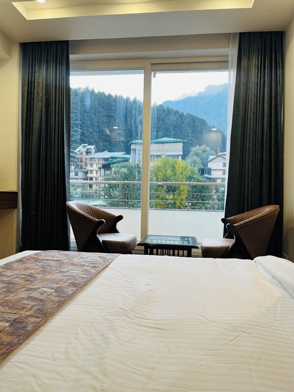 A hotel room with a large bed, two chairs, a small table, and a balcony with a view of buildings and mountains.