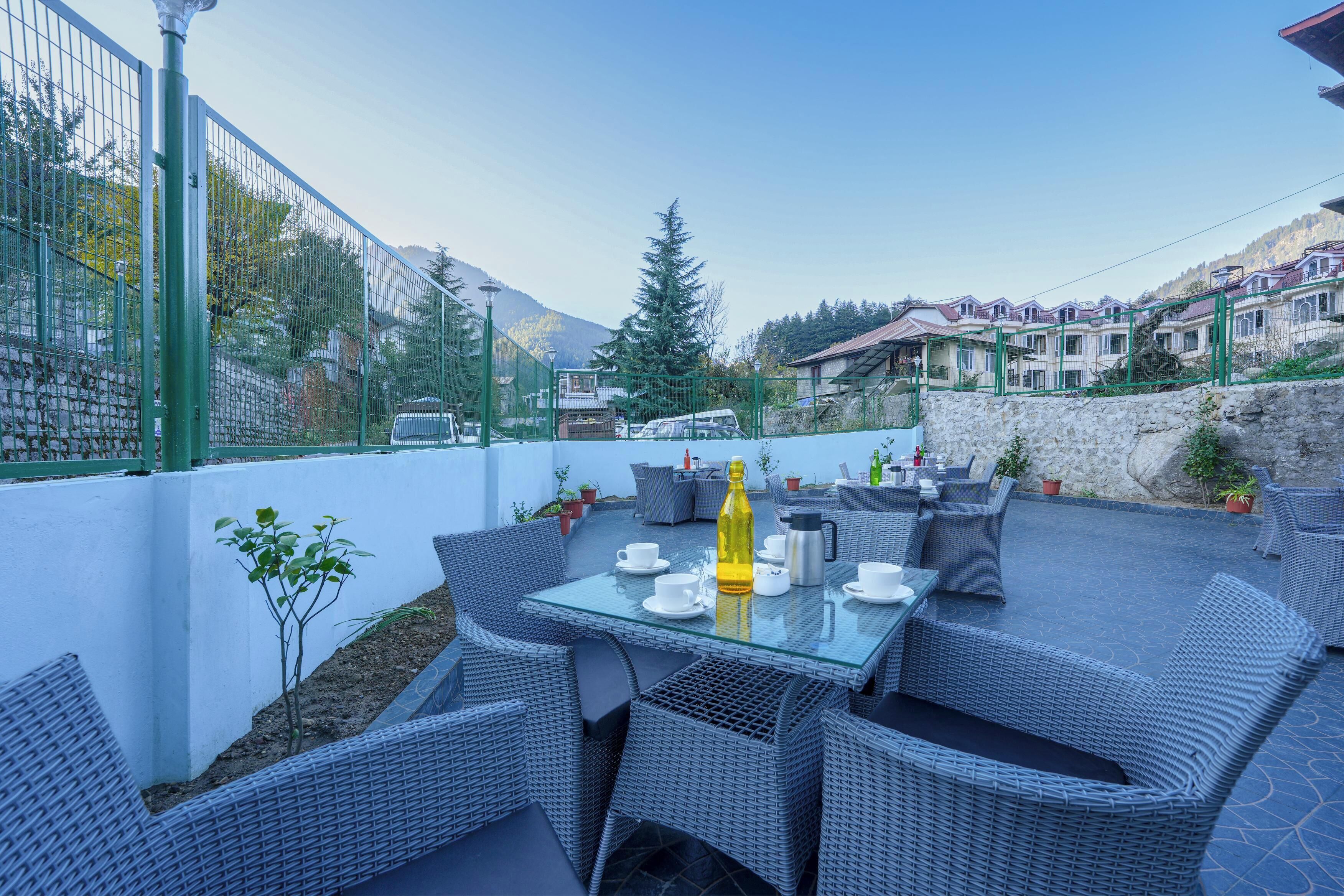 An outdoor patio with wicker furniture and a glass table, surrounded by a stone wall and a metal fence.