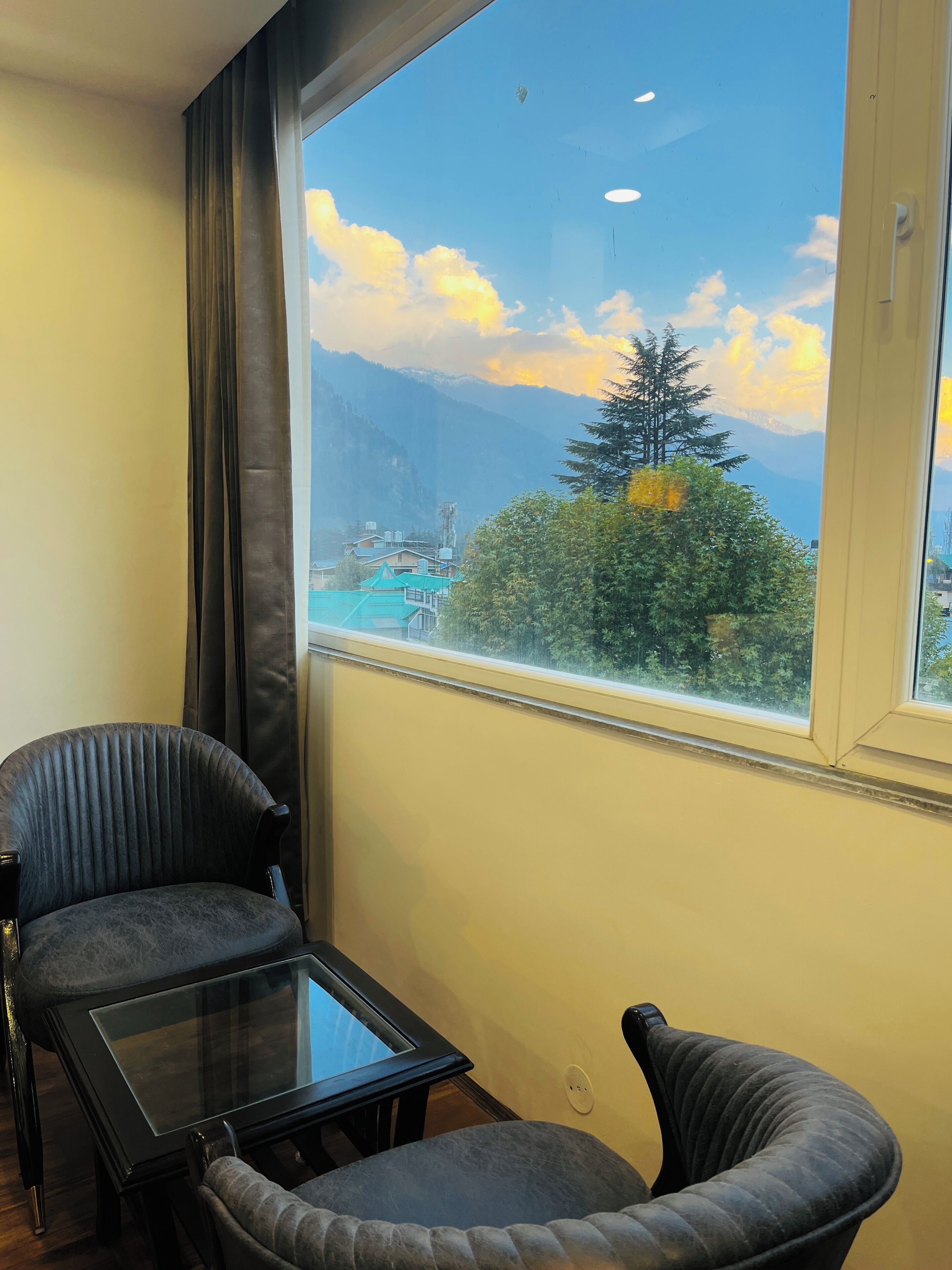 A room with a large window offering a mountain view, two upholstered chairs, and a glass-top table.