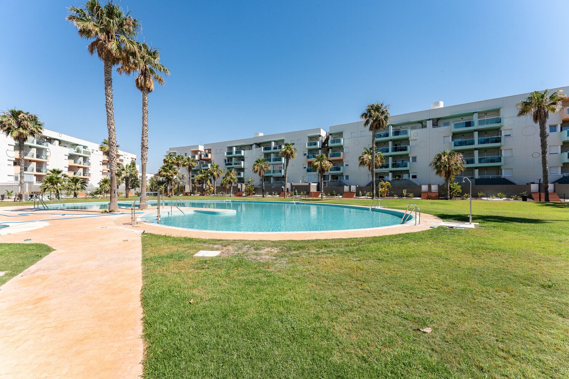 Un área de piscina con cielo despejado, palmeras y un edificio moderno con balcones.