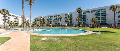 Un área de piscina con cielo despejado, palmeras y un edificio moderno con balcones.