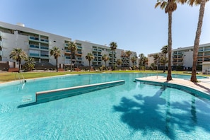 A swimming pool with clear blue water, surrounded by palm trees and a multi-story building with balconies.