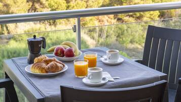 Een balkon met een tafel waarop croissants, fruit, sap en koffiekopjes staan, met uitzicht op een bebost gebied.