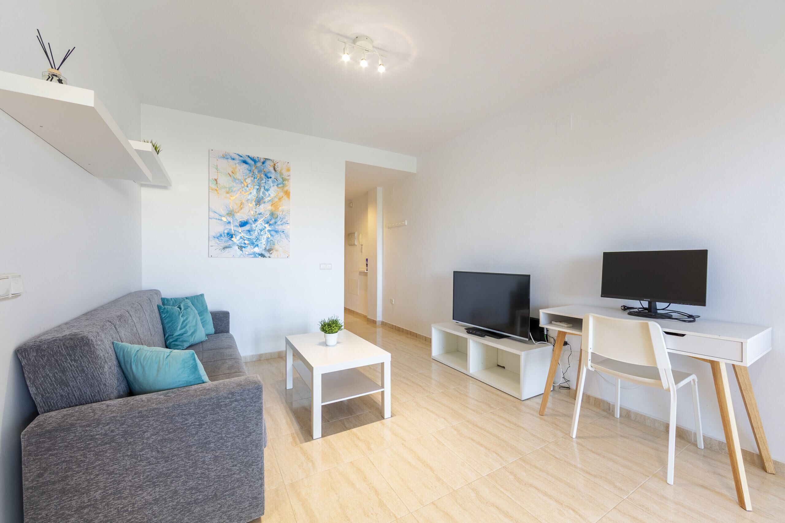 Una sala de estar moderna con un sofá gris, una mesa de centro blanca, un televisor y un escritorio con una silla.