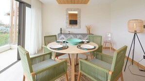 A dining area with a round table, green upholstered chairs, a decorative mirror above, and a view through a large window.