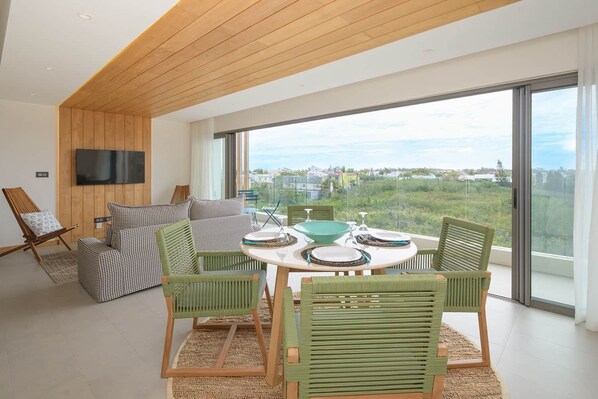 A modern living room with a wooden ceiling, a large window with a city view, a round dining table with chairs, and a TV mounted on the wall.