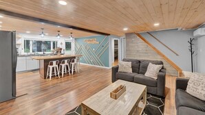 A spacious living area with a wooden ceiling, a kitchen with a bar, and a gray sofa with patterned cushions.