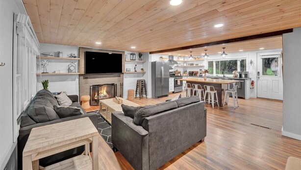 A modern living room with a fireplace, grey sofas, a wooden coffee table, and a kitchen area with a refrigerator and island.