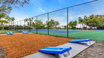 Een tennisbaan met een hek, blauwe bankjes en een boom op de achtergrond.