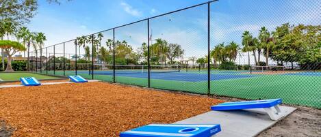 Una cancha de tenis con valla, bancos azules y un árbol al fondo.