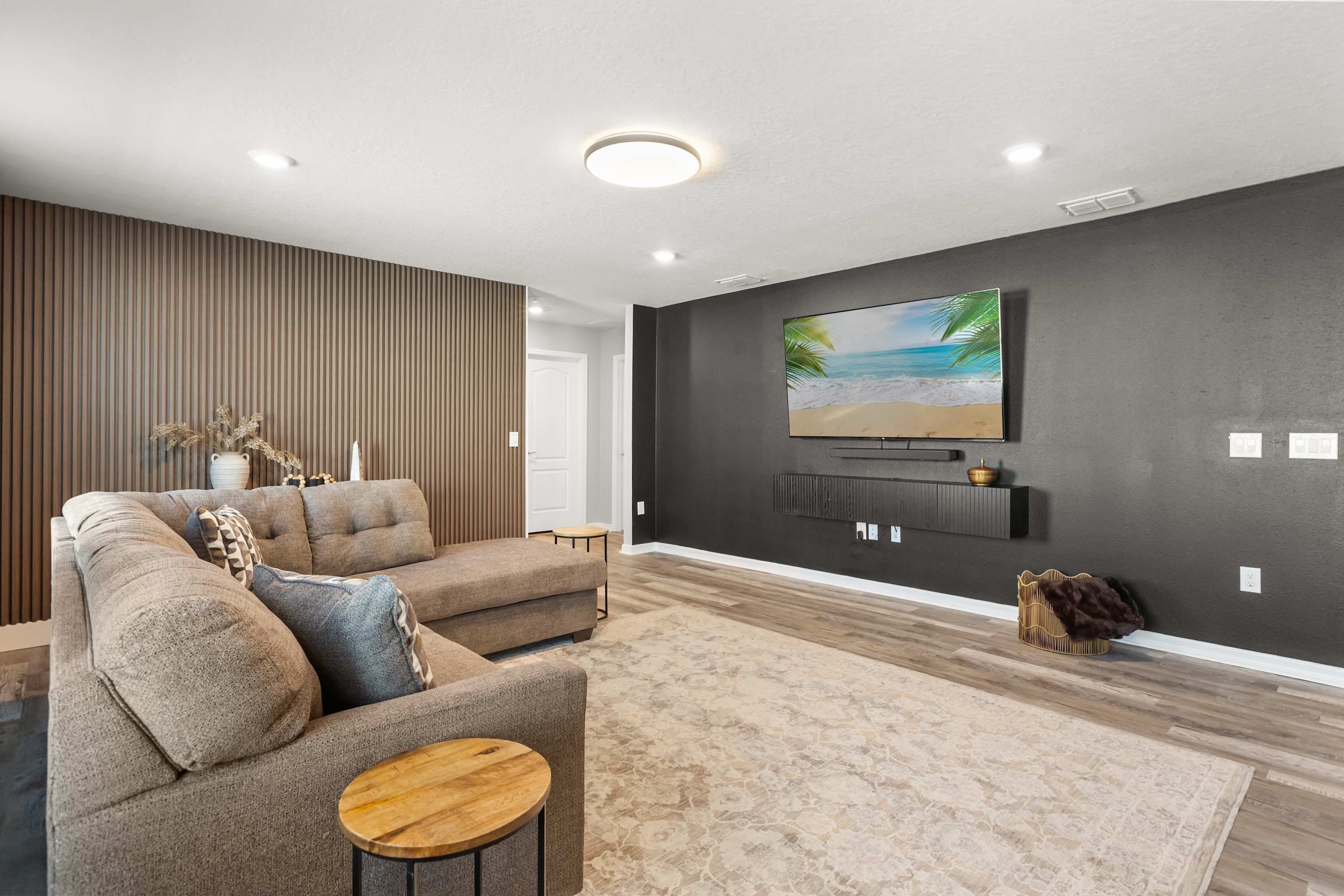 Un salon moderne avec un canapé d’angle, une table basse ronde en bois, un grand téléviseur à écran plat fixé à un mur foncé et un tapis à motifs sur le plancher en bois.