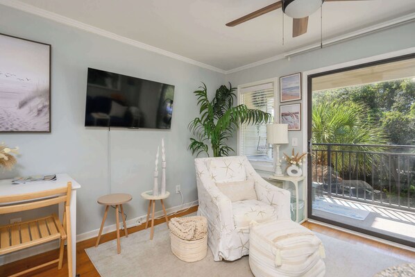 A room with a wooden desk, a white chair, a large plant, and a balcony with a view of greenery.