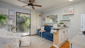 A living room with a sofa, a small table, a ceiling fan, a kitchen area with cabinets, and a balcony with a view of greenery. - Island Condo for 6 w Communal Pool - Perfect Spot! (Hilton Head Island)