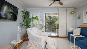 A living room with a ceiling fan, a TV, a sofa, a chair, a table, and a large window. - Island Condo for 6 w Communal Pool - Perfect Spot! (Hilton Head Island)