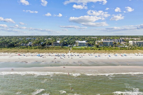 A beach with a hotel in the background, a sandy shore, and waves. - Island Condo for 6 w Communal Pool - Perfect Spot! (Hilton Head Island)