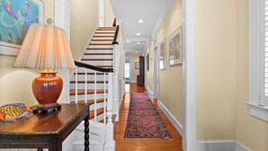 Un couloir avec un escalier en bois, un tapis rouge à motifs et une table sur laquelle se trouvent une lampe et des objets décoratifs.