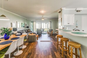 Amplia sala de estar con mesa de comedor de madera, sillas blancas y sofá. Se observa una zona de cocina con encimera blanca y taburetes de bar.
