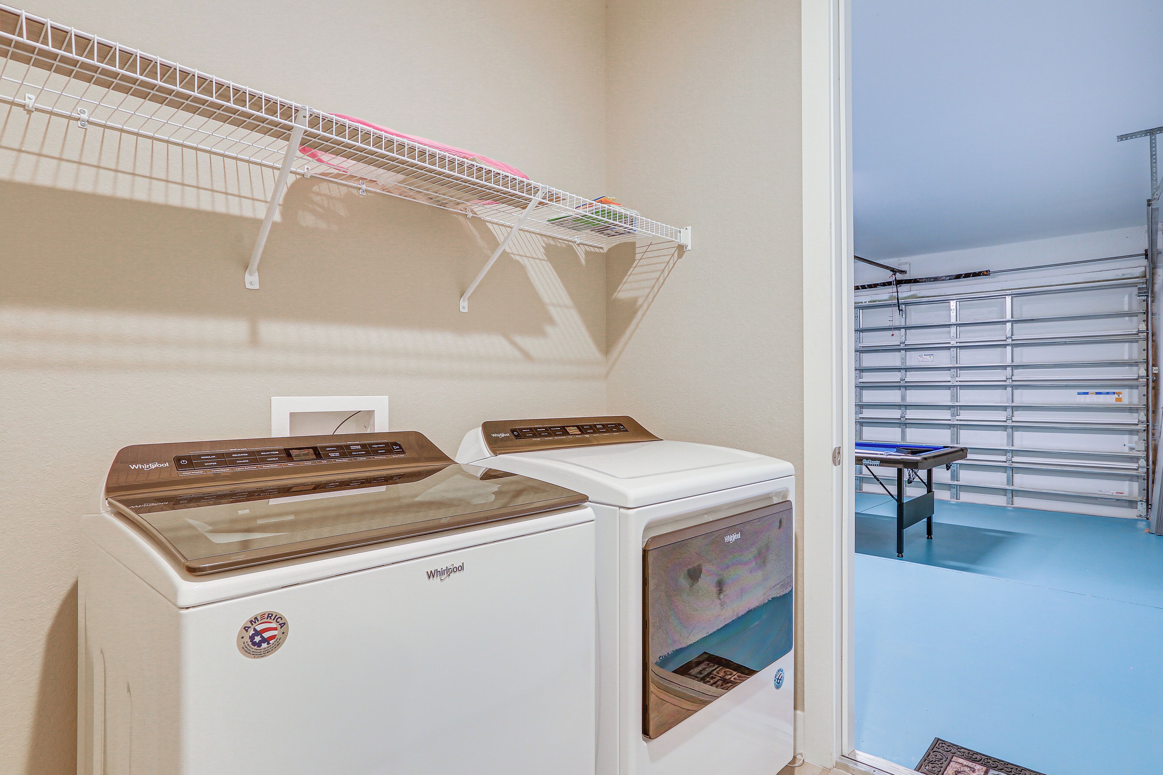 A laundry area with two Whirlpool dryers, a metal shelf with laundry baskets, and a view of a garage with a blue floor.