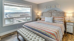 A bedroom with a large window, a bed with a patterned bedspread, two bedside lamps, and a bench at the foot of the bed.