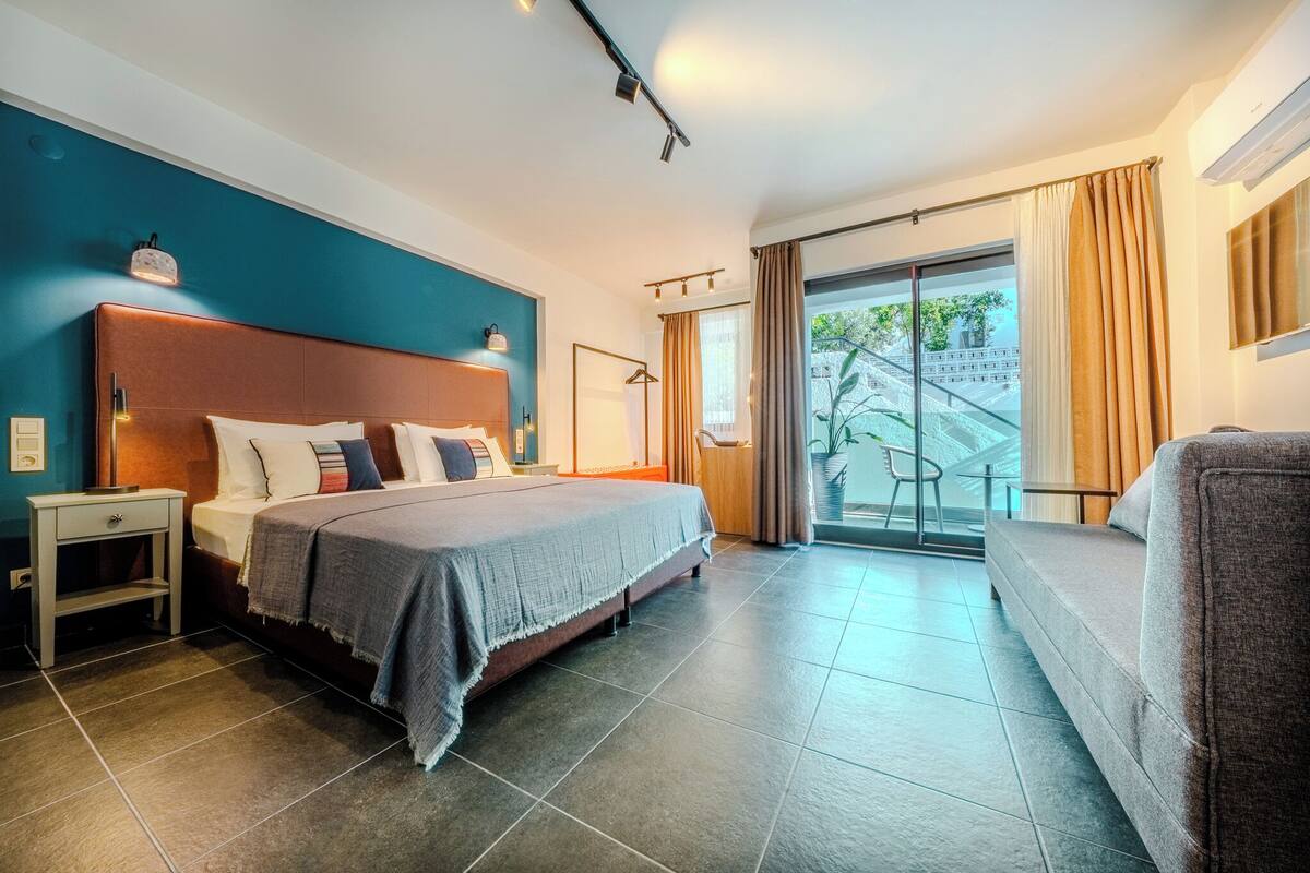 Een moderne hotelkamer met een groot bed, een bank, een klein tafeltje en uitzicht op een trap die naar buiten leidt.