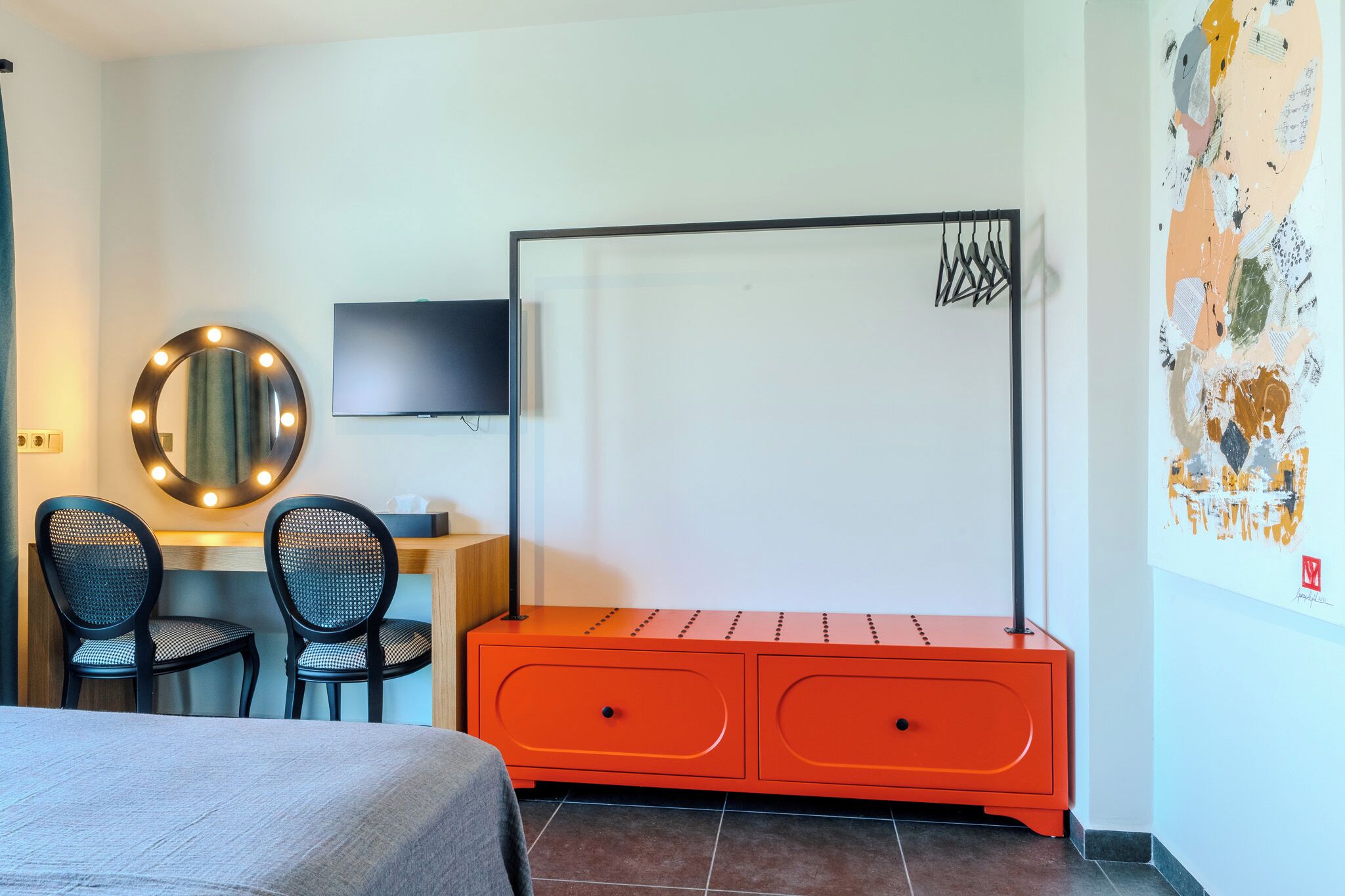 A hotel room with a bed, two chairs, a desk with a mirror and lights, a TV, an orange cabinet, and a wall with artwork.
