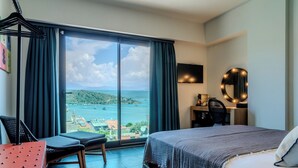 A hotel room with a large window offering a view of a coastal area, a bed with a grey blanket, a desk with a chair, and a round mirror with lights.