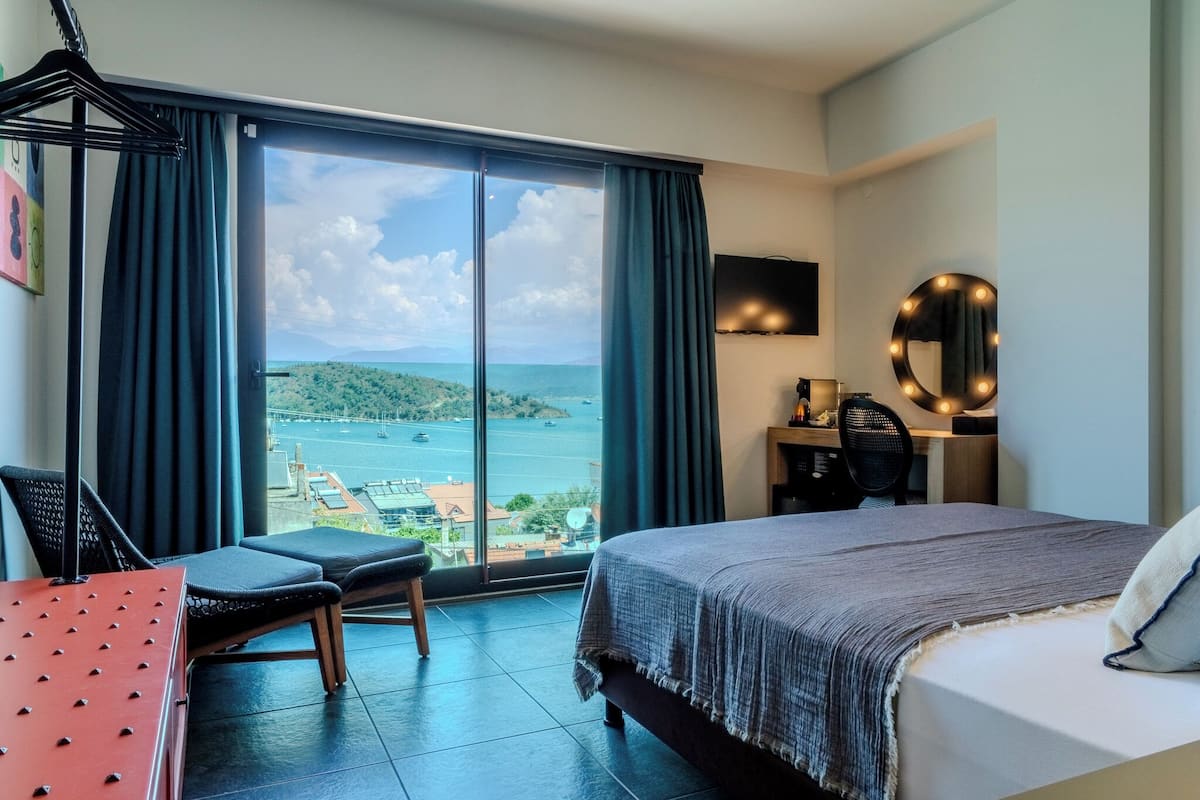 Hotelkamer met een groot raam en uitzicht op de kust, een bed met een grijze deken, een bureau met stoel, en een ronde spiegel met verlichting.