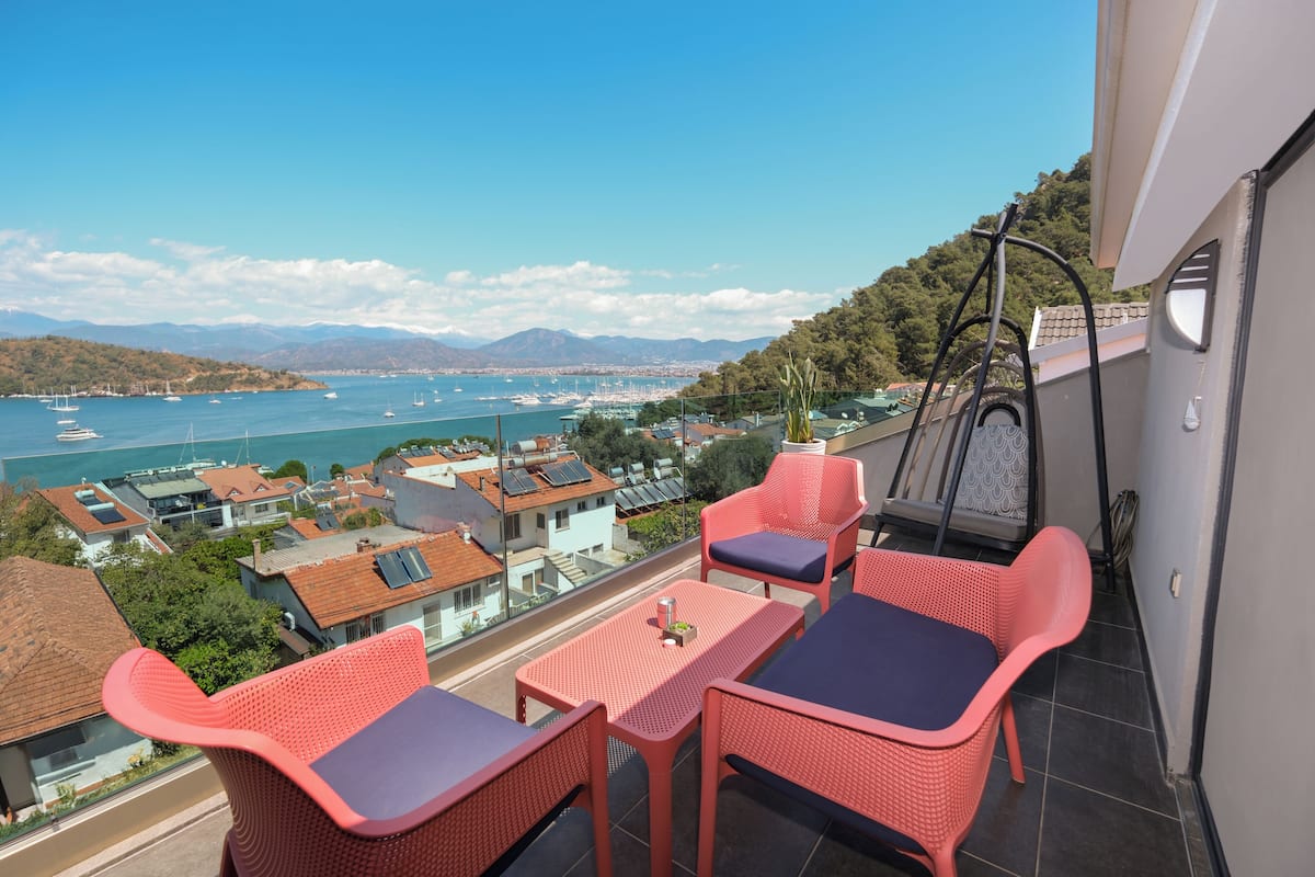 Een balkon met rode rieten meubels, uitzicht op zee en gebouwen op de achtergrond.