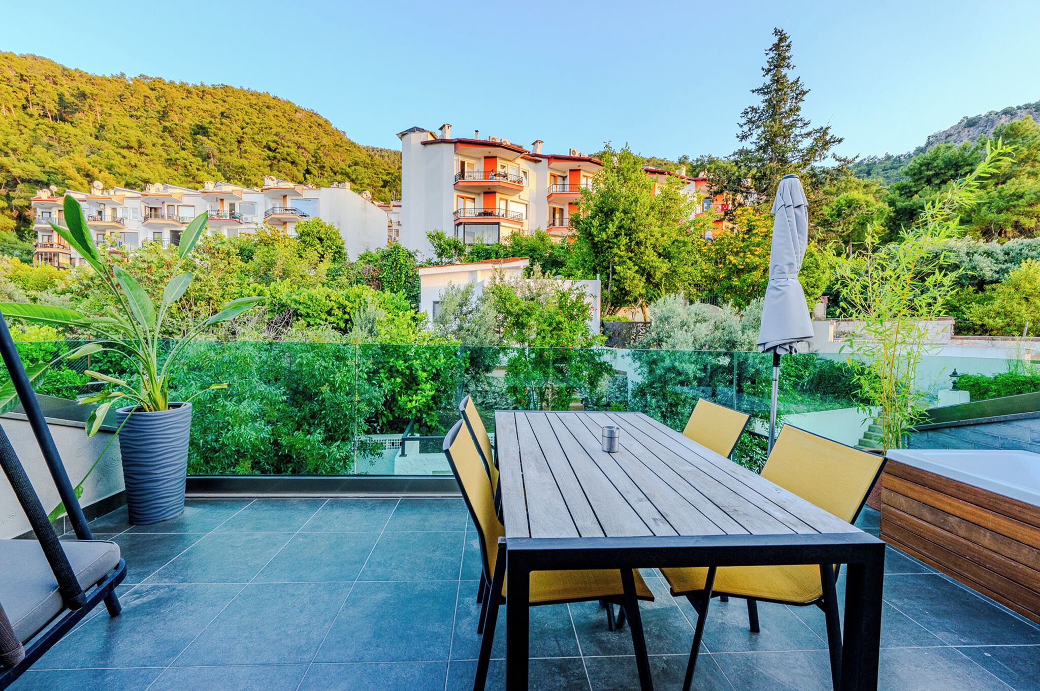 A terrace with a table and chairs, a potted plant, and a view of buildings and greenery.